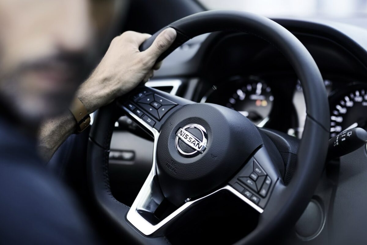A driver's hand on the wheel of a Nissan, the badge and dashboard in view